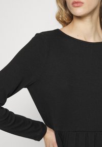 Schwarzes Langarmkleid mit rundem Ausschnitt und geraffter Taille, aus weichem, strukturiertem Stoff.