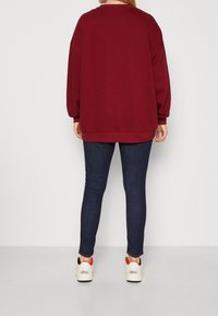 Sweatshirt oversize bourgogne avec épaules tombantes, associé à un jean slim-fit bleu foncé et des baskets blanches avec des accents rouges.