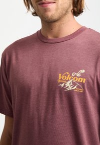 Weinrote Baumwoll-T-Shirt mit einem gelben Grafiklogo auf der Brust. Kurze Ärmel und klassischer Rundhalsausschnitt mit weicher Textur.