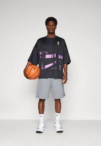 Jaunas vyras stovi, vienoje rankoje laikydamas krepšinį, vilkėdamas juodą per didelį NBA marškinėlį, pilkas šortus, baltas kojines ir baltus Nike sportbačius.