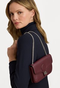 Burgundy leather handbag with a chain strap, featuring a flap closure and embossed logo detail on the front, carried on a shoulder.