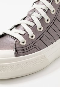 Metallic lila sneaker med vita snören, vit gummisula och detaljerad sömnad på en vit bakgrund.