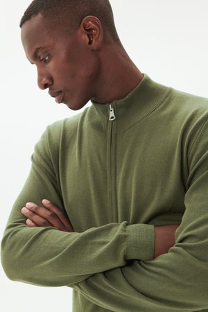 Suéter de punto con cremallera en verde oscuro, con cuello alto y puños acanalados. Textura suave, corte relajado y un ligero brillo.