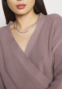 Suéter color malva claro con diseño de escote en V, textura acanalada y collares de cadenas plateadas en capas. Primer plano del torso superior.