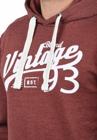 Bordov�ý mikinový sveter vyrobený z jemnej tkaniny s bielymi šnúrkami. Na prednej strane sa nachádzajú veľké biele písmo "Vintage" a grafický detail "EST. 93".