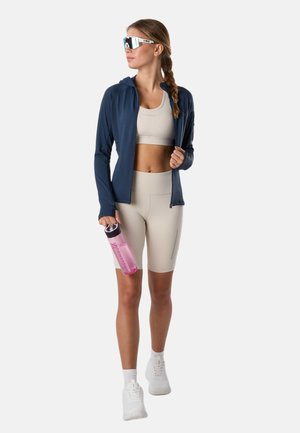 Frau in beigefarbenem Sport-BH und Shorts mit marineblauer Jacke, weißen Turnschuhen, Sonnenbrille und einer rosa Wasserflasche in der Hand, stehend vor weißem Hintergrund.