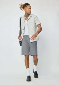 Chemise légère grise à manches courtes et texturée sur un débardeur blanc, associée à un short gris. Des mocassins noirs et des chaussettes blanches viennent compléter le look.