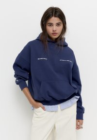 Marineblauwe hoodie met een grote voorkantzak, geborduurde witte tekst en een gelaagde gestreepte shirt aan de onderkant. Zachte stof en een oversized pasvorm.