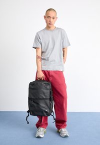 Svart ryggsäck med flera fack, bärs av en person i en ljusgrå t-shirt och röda byxor, placerad mot en neutral bakgrund.