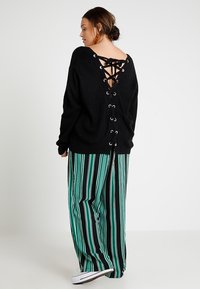 Zwarte gebreide oversized trui met een vetersluiting op de achterkant. Gecombineerd met groen en zwart gestreepte wijde broek en witte schoenen.