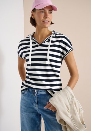 Femme portant un sweat à capuche manches courtes rayé noir et blanc, casquette rose, jean bleu, tenant une veste beige dans une main sur fond neutre.