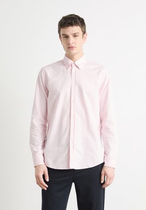 Chemise boutonnée en coton rose clair avec des manches longues, un col cranté et une texture subtile ; associée à un pantalon sombre pour créer un contraste.