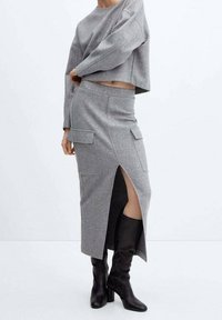 Pull gris court et jupe longue assortie avec fente latérale et deux poches avant ; porté avec des bottes noires montantes. Texture de tissu lisse.