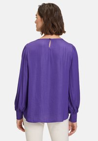Lilla bluse med lange, puffede ærmer; glat tekstur; nøglehulslukning bagpå; afslappet pasform, velegnet til forskellige anledninger.