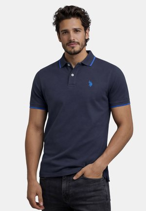 Mann mit Bart, der ein dunkelblaues Poloshirt mit blauem Rand und Logo trägt, posiert mit einer Hand in der Tasche vor einfachem Hintergrund.