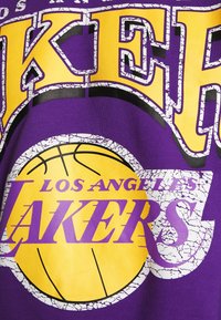 T-shirt en coton violet avec un grand texte jaune et blanc indiquant "LOS ANGELES LAKERS", avec un graphique de ballon de basket fissuré au centre.