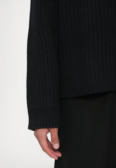 Mano rilassata accanto al corpo, indossando un maglione a coste nero con polsini e pantaloni neri su sfondo bianco.