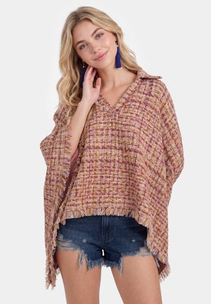 Donna bionda che indossa un poncho rosa e beige con trama, con frange e orecchini blu con nappine, abbinato a shorts di jeans consumati.