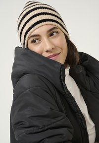 Doudoune noire avec un col haut, fermeture éclair, et un bonnet en tricot à rayures beige et noires. Texture lisse et design léger.