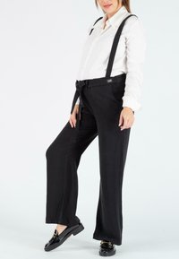 Pantalon noir avec bretelles, design à jambes larges, texture du tissu visible, associé à une chemise blanche et des mocassins noirs, avec un nœud à la taille.