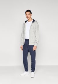 Sweat à capuche gris clair zippé avec cordons, associé à un pantalon de jogging bleu marine. T-shirt blanc en dessous, et baskets blanches complètent la tenue.