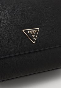 Musta teksturoitu käsilaukku, jossa on kultainen kolmion muotoinen logo-plate, jossa lukee "GUESS USA" ja kysymysmerkki symboli. Saumadetailit parantavat sen muotoilua.