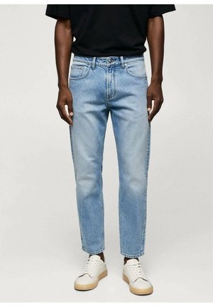 Mężczyzna ubrany w jasnoniebieskie proste dżinsy, czarną koszulę i białe trampki, stojący na jednolitym tle.