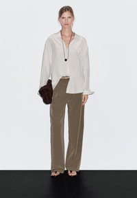 Soft, cream blouse with a collar, loose fit; tan, wide-leg trousers; brown clutch bag; long black necklace with a round pendant.