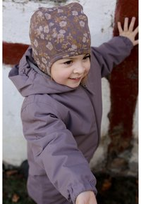 Lila wasserdichte Jacke mit Kapuze. Gemusteter brauner Blumenhut. Kind streckt die Hand aus, Hände an einer strukturierten weißen und roten Wand.