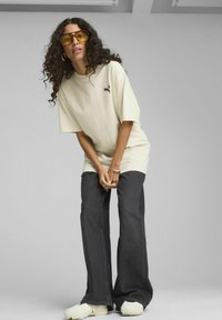 T-shirt beige oversize con una piccola grafica, abbinato a jeans scuri a zampa d'elefante e scarpe bianche con texture. Modello che indossa occhiali da sole gialli.