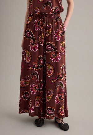Bruine maxirok met kleurrijke roze, oranje en groene paisleypatronen, elastische taille, zijzakken, gedragen met zwarte gevlochten platte schoenen op een neutrale achtergrond.