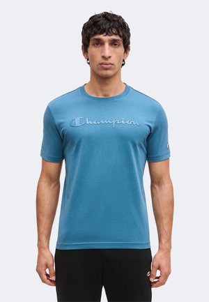 Mann trägt blaues kurzärmeliges T-Shirt mit "Champion"-Logo auf der Brust und am linken Ärmel, kombiniert mit schwarzen Hosen vor einem schlichten Hintergrund.