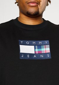 Černá mikina s obdélníkovou vyšívanou nášivkou. Nášivka má námořnicky modrý okraj s nápisem "TOMMY JEANS" a v centru károvaný vzor.