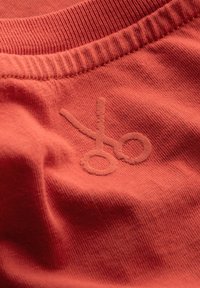 Koraalstof met een getextureerd, geborduurd schaarlogo. De stevige stiksels langs de halslijn zorgen voor duurzaamheid.