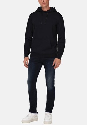Zwarte hoodie met een kangoeroezak, geribbelde boorden en een trekkoord. Gecombineerd met donkerblauwe slim-fit jeans en witte sneakers.