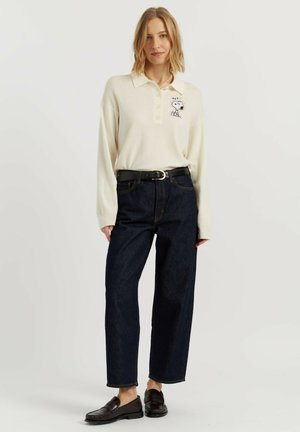 Vrouw draagt een lange mouwen crème poloshirt met Snoopy borduurwerk, donkere hoog getailleerde jeans, zwarte riem en zwarte loafers, staand tegen een witte achtergrond.