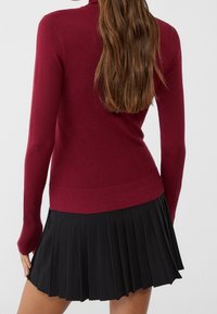 Pull en maille bordeaux à manches longues avec ourlet côtelé, associé à une jupe plissée noire présentant des plis nets et une texture lisse.