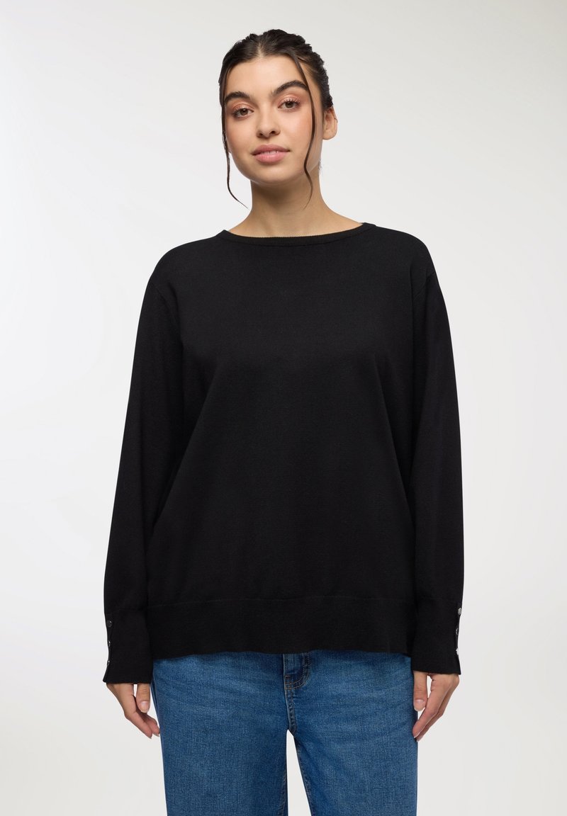 Czarny sweter dzianinowy z długimi rękawami, okrągłym dekoltem i guzikami na mankietach. W zestawieniu z niebieskimi dżinsami. Gładka tekstura.