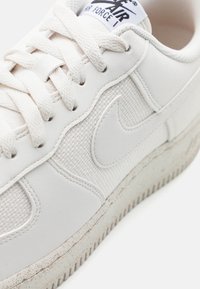 White leather sneaker with a textured side panel, flat laces, and a rubber sole. Features a prominent logo on the tongue and subtle dirt marks.