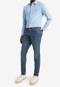 Camisa azul claro abotonada con un logo en el pecho, combinada con jeans azul oscuro y zapatillas blancas. La tela parece suave con un ajuste casual.