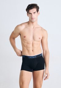 Homem sem camisa com cabelo curto e crespo, vestindo boxers Lacoste pretos com uma faixa azul, em pé contra um fundo claro e simples.