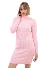 Žena s krátkými blond vlasy, oblečená v růžových svetrkách s copánkovým vzorem a rolákem, pózuje s jednou rukou u krku.
