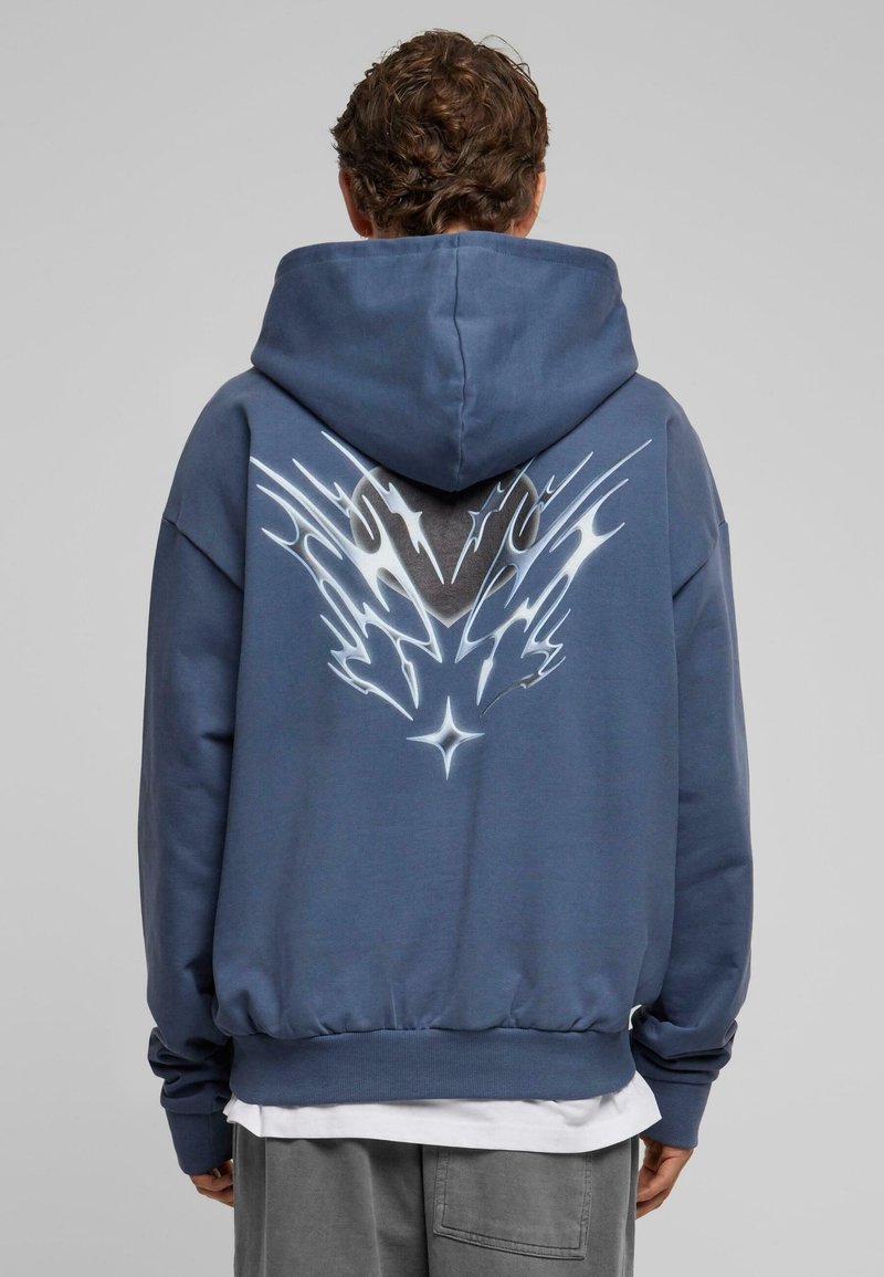 Homme portant un sweat à capuche bleu avec un motif abstrait blanc en forme de flamme dans le dos, debout devant un fond gris uni.