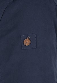 Tela azul marino con un pequeño parche de cuadrados cosido que presenta un emblema redondo de bronce grabado con un ancla y estrellas.