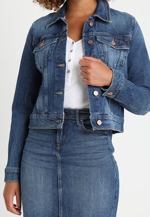 Veste en denim bleu profond avec une coupe ajustée, un devant boutonné, deux poches poitrine et des détails en métal, associée à un haut blanc.