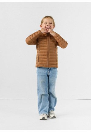 Jong kind draagt een bruine gewatteerde jas, lichtblauwe spijkerbroek en witte sneakers, glimlacht en met de rits van de jas bezig.