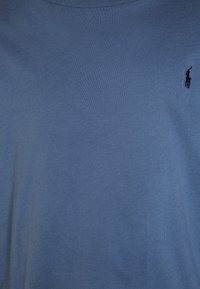 Blå bomullströja med korta ärmar och rund hals. Har en liten svart broderad logotyp på vänster bröstområde. Mjuk textur.