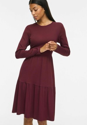Femme aux longs cheveux foncés portant une robe bordeaux à manches longues, longueur genoux, debout, les mains jointes et regardant vers le bas.