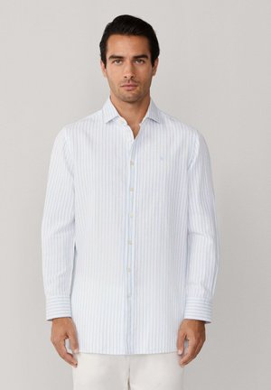 Homme de face portant une chemise blanche à manches longues avec de fines rayures verticales bleues, debout devant un fond uni clair.