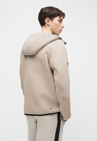 Jonge man draagt een beige gewatteerde jas met capuchon en lichte broek met zwarte zijstrepen, staand zijwaarts met handen in de zakken.